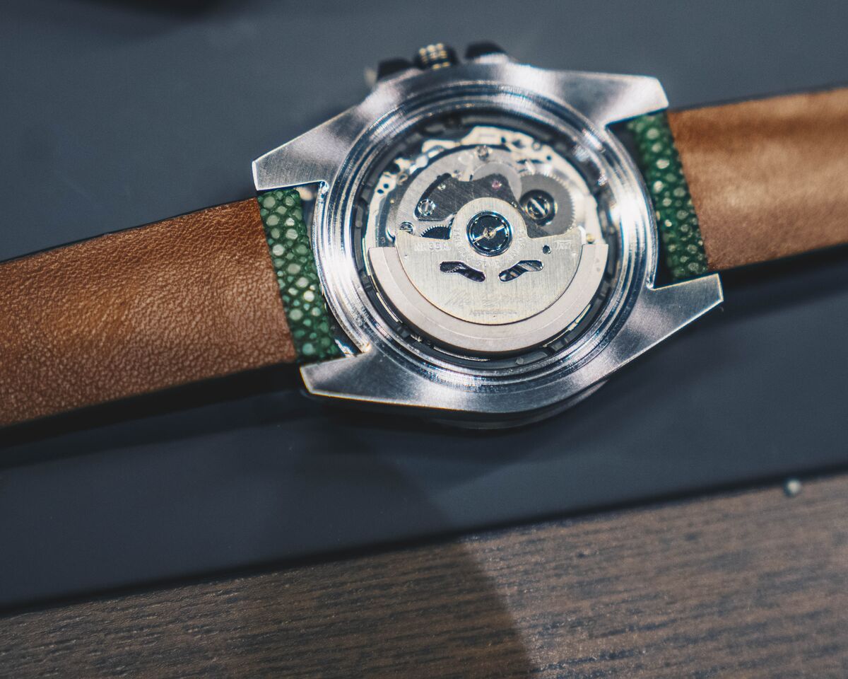 Caseback macro of a watch on a green and tan strap, with the movement visible through the back.
